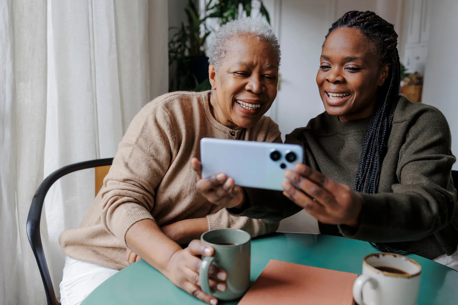 A caregiver and an older woman laughing over a phone at the kitchen table