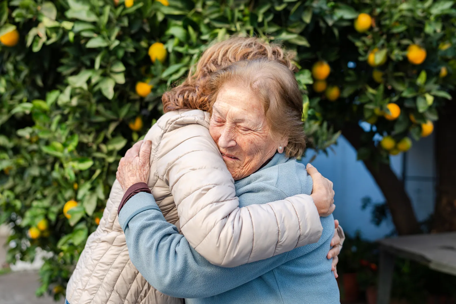 A caregiver and an older woman hugging under an orange tree